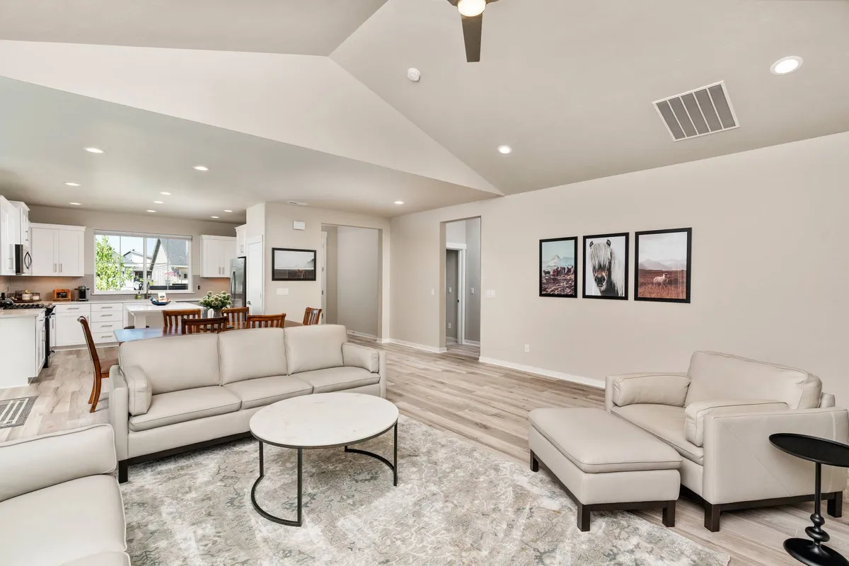 An expansive, sunlit open-concept interior featuring a comfortable sofa set and coffee table in the foreground, with a stylish kitchen area visible in the background. Wall artwork and custom décor add a contemporary, inviting flair to the space, which is clearly designed for social gatherings and family connection.
