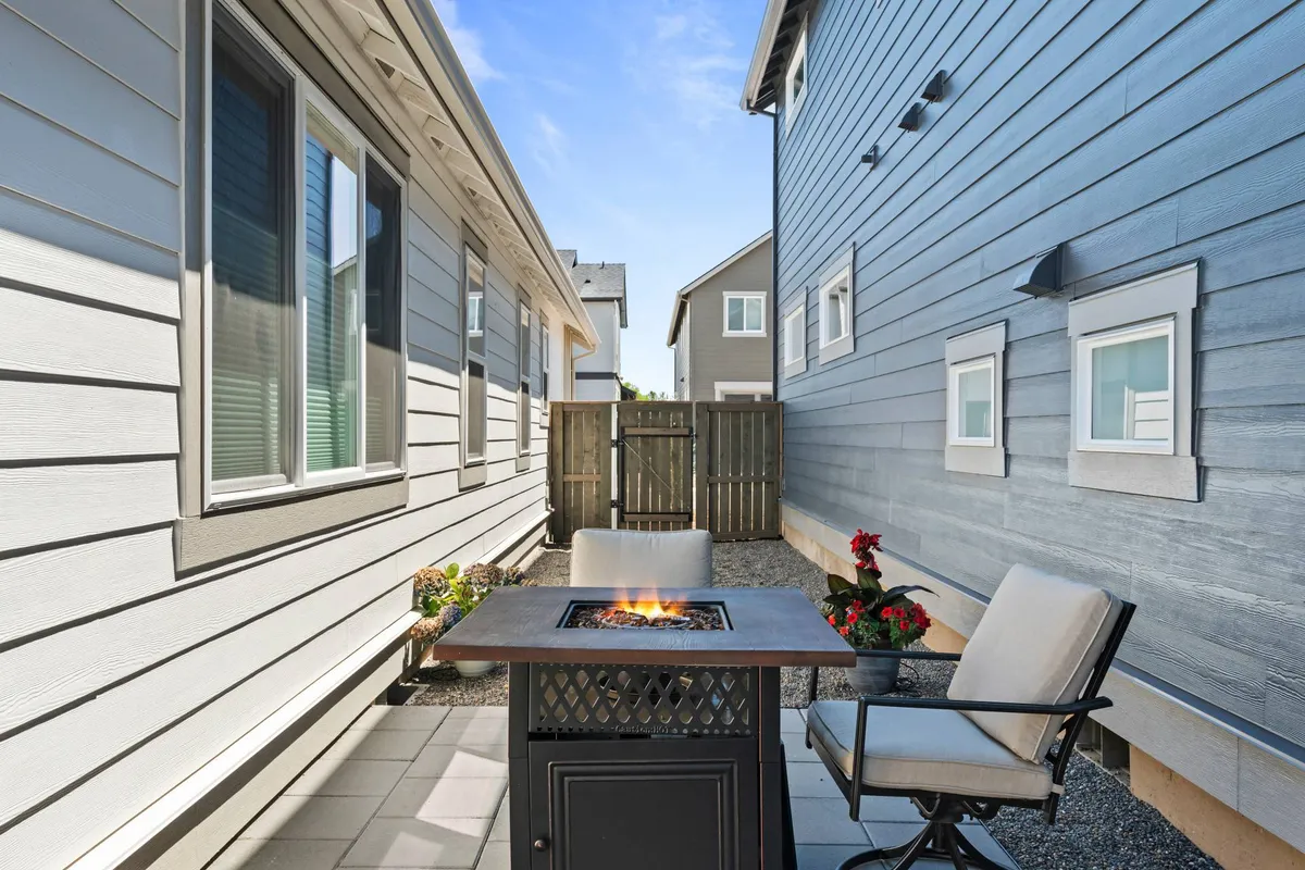 An inviting exterior shot of a private patio complete with a cozy fire pit, ample seating, and vibrant flower pots. The scene suggests warm evenings spent outdoors, ideal for gathering and unwinding as a family or with friends.