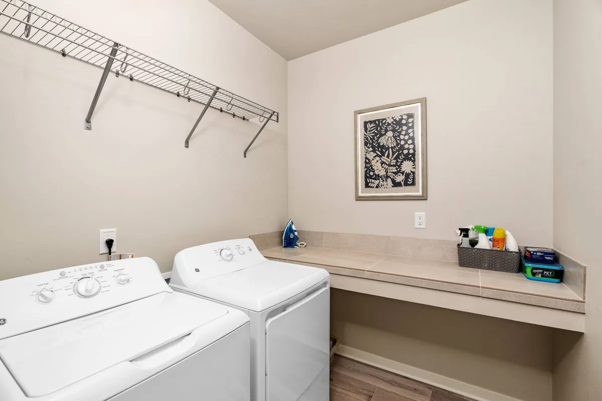 This image showcases a well-organized laundry room featuring a washing machine, drying machine, countertop for folding, wall art, and additional shelving for storage and supplies. The space is clean, functional, and designed to make laundry tasks efficient and pleasant.