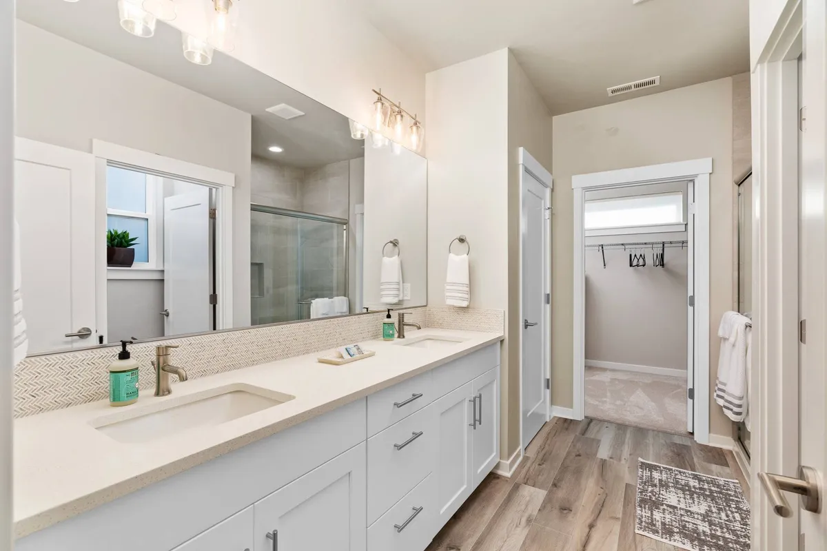 A well-lit bathroom showcasing a spacious double vanity with two sinks, a large mirror, light fixtures, and a view toward the shower area. The clean design and fresh ambiance evoke a spa-like feel, perfect for self-care routines.