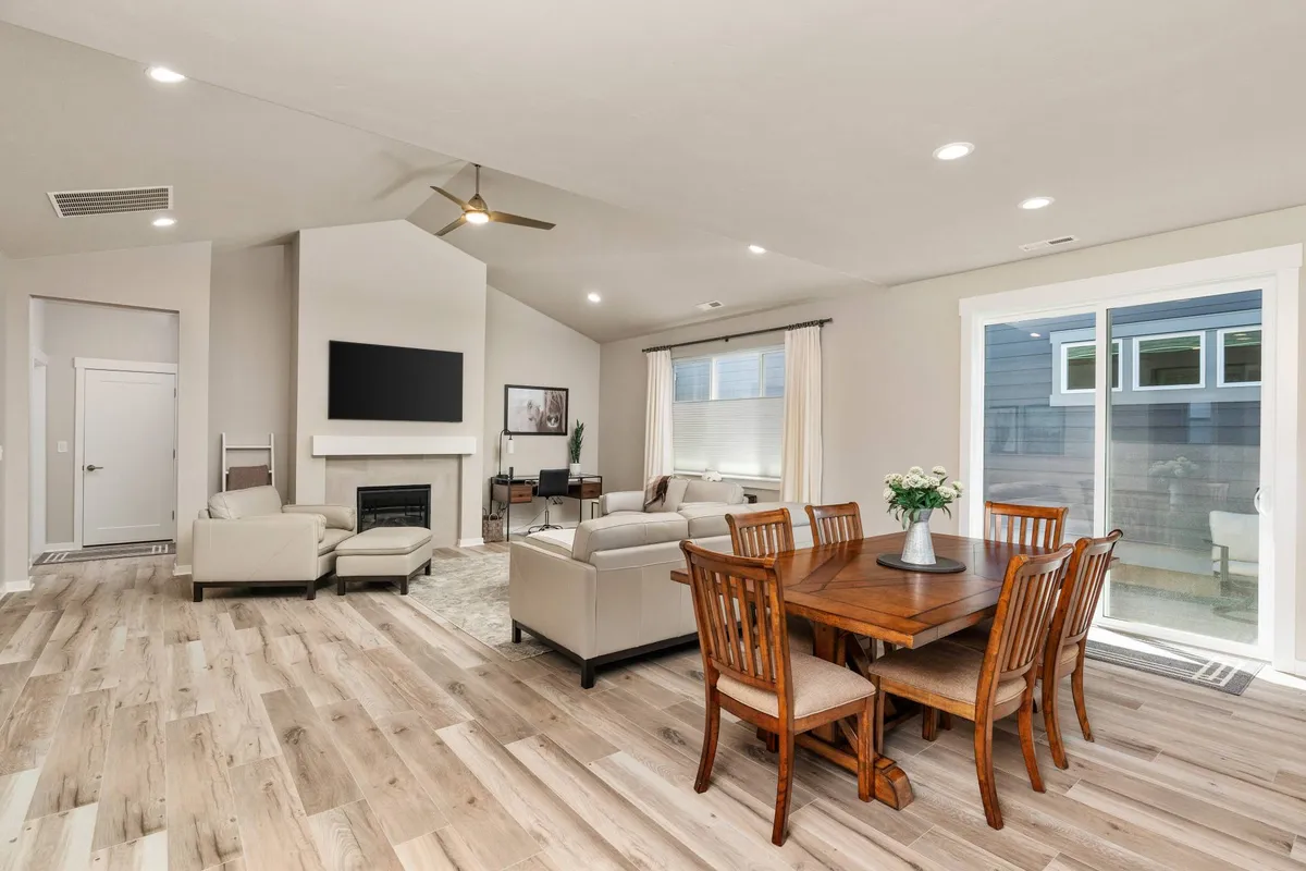 A cozy interior shot showing a dining table set for meals, a sofa for comfortable lounging, and a fireplace as the room's focal point. Large windows bathe the space in natural light, enhancing the inviting and communal atmosphere.