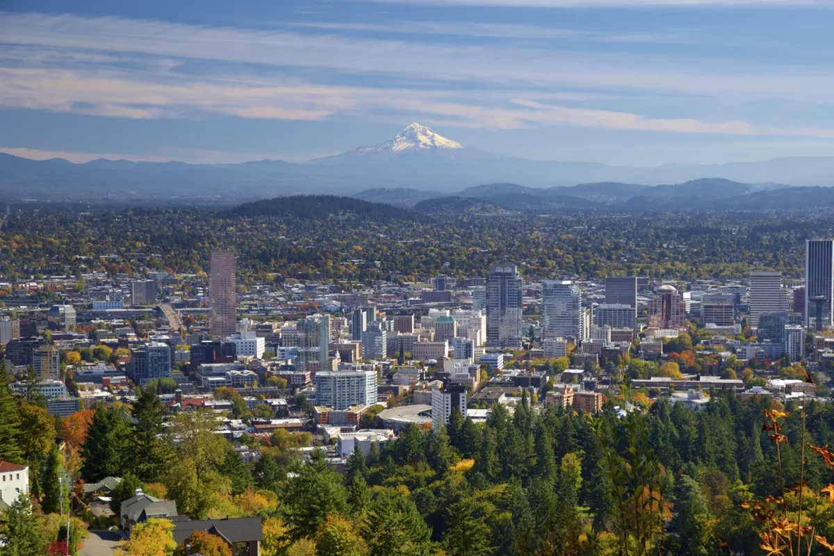 A panoramic view featuring Mount Hood rising above the city skyline, framed by green trees and autumn foliage. The image conveys the natural beauty easily accessible from the location and a sense of tranquility at the doorstep of urban life.