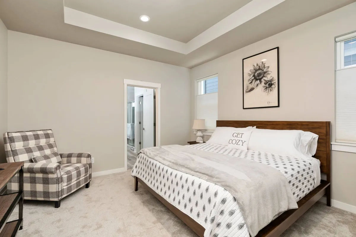 A cozy bedroom scene featuring a plush bed adorned with decorative pillows, a plaid armchair in the corner, framed wall art, and a bedside lamp creating a serene ambiance. Natural light filters softly through the window, enhancing the inviting atmosphere.